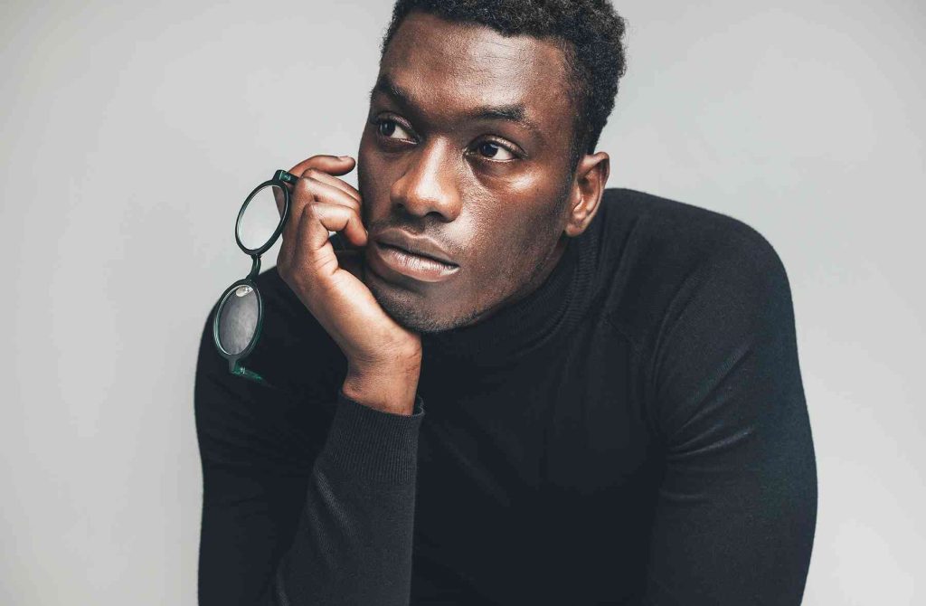 A contemplative man holding glasses, gazing thoughtfully, dressed in a black turtleneck.
