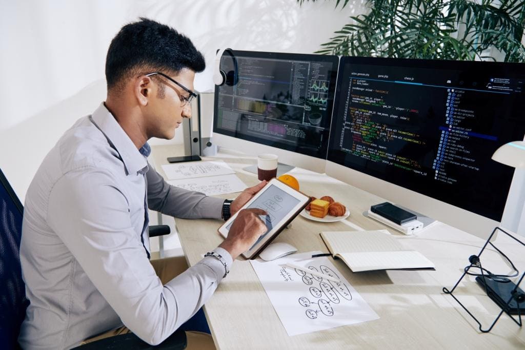 Young man analyzing code on dual monitors in a tech workspace.